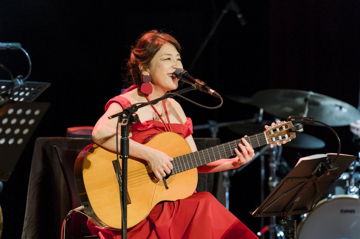 Bossa nova singer Lisa Ono performs live in a red dress, singing into a microphone while playing an acoustic guitar on stage.